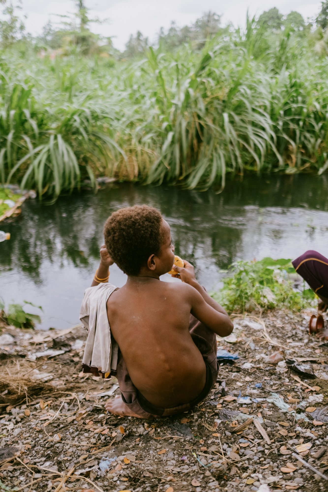 A small child sits alone on the riverbank, watching the water
